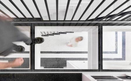 top-down angle of a man walking in his home on glass flooring. The man's wife can be seen walking on the lower floor.